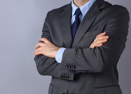 A man in a dark suit with his arms crossed. A wristwatch is visible.