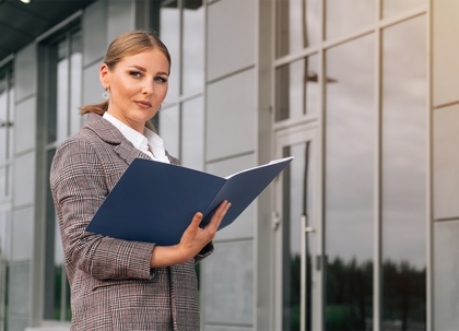 A woman in a business suit holds a folder, standing in front of a house.