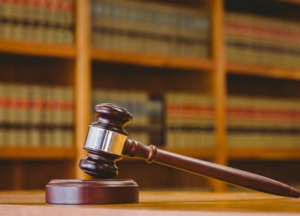 A wooden gavel rests on a table, in front of a bookcase filled with law books.