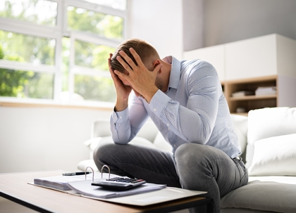 A man in a suit is hunched over blueprints, his head in his hands, looking distressed.