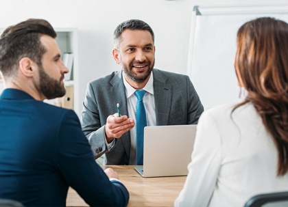 A man in a gray suit is seated, likely in a meeting, with other people present.