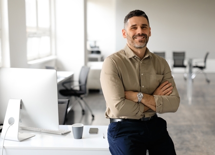 A man in a light brown shirt, arms crossed, stands at a white desk with computers and a mug. He's smiling.