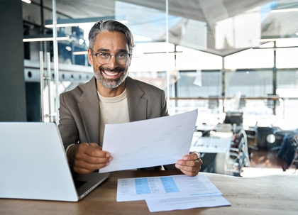 A man in a beige jacket and glasses, smiling, holds papers over a laptop on a desk in an office.