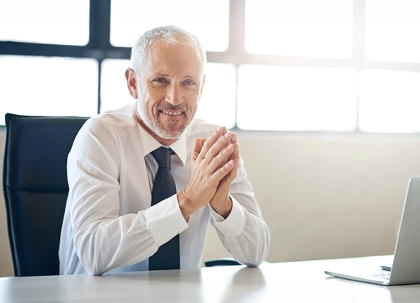 A man in a business suit is seated at a desk, smiling.