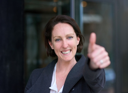 A woman with brown hair is smiling and giving a thumbs-up.