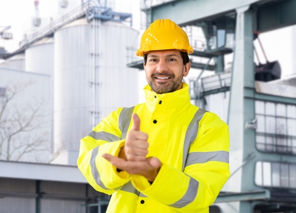 A man in a yellow safety vest and hard hat, giving a thumbs-up, in front of industrial buildings.