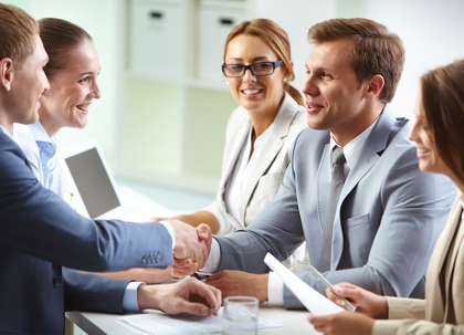 Several business people are seated around a table, shaking hands and talking.