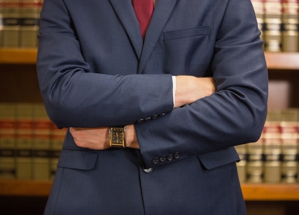 A man in a navy suit, arms crossed, stands in front of a bookcase filled with law books.