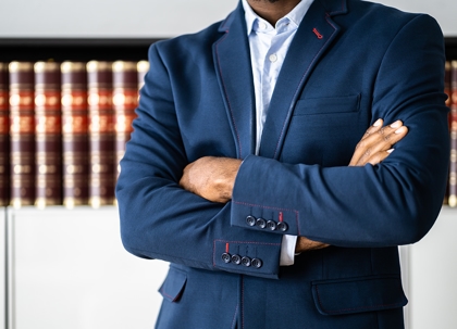 A man in a dark suit, arms crossed, standing in front of bookshelves and a table.