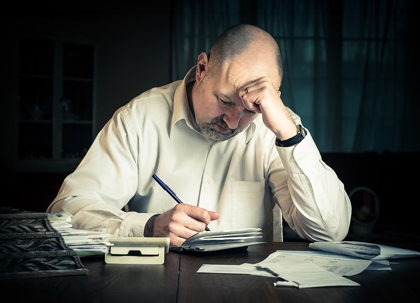 A man sits at a table, surrounded by bills and a calculator, looking stressed.