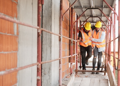 Two construction workers, one helping the other, walk along a scaffolding walkway.