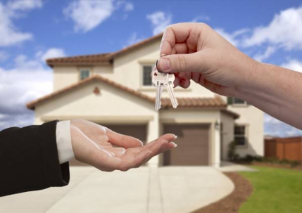 Two hands exchanging house keys in front of a house.