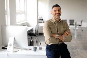 A man in a light brown shirt, arms crossed, stands at a white desk with computers and a mug. He's smiling.