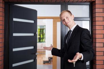 A man in a suit stands at an open doorway, gesturing, with keys in his hand.
